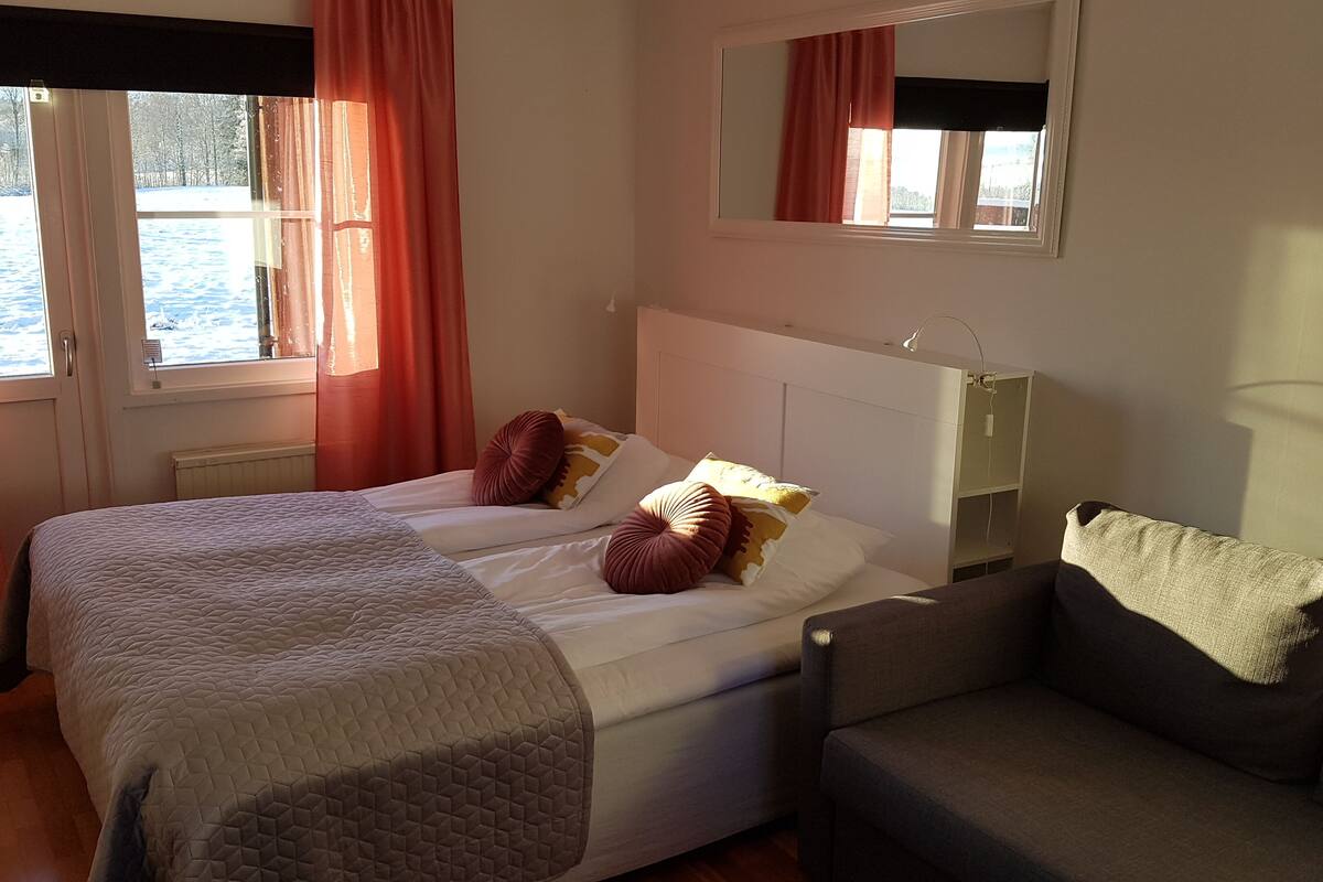 A bedroom with a bed, a bedside table, a lamp, a window with curtains, and a view of the outdoors.