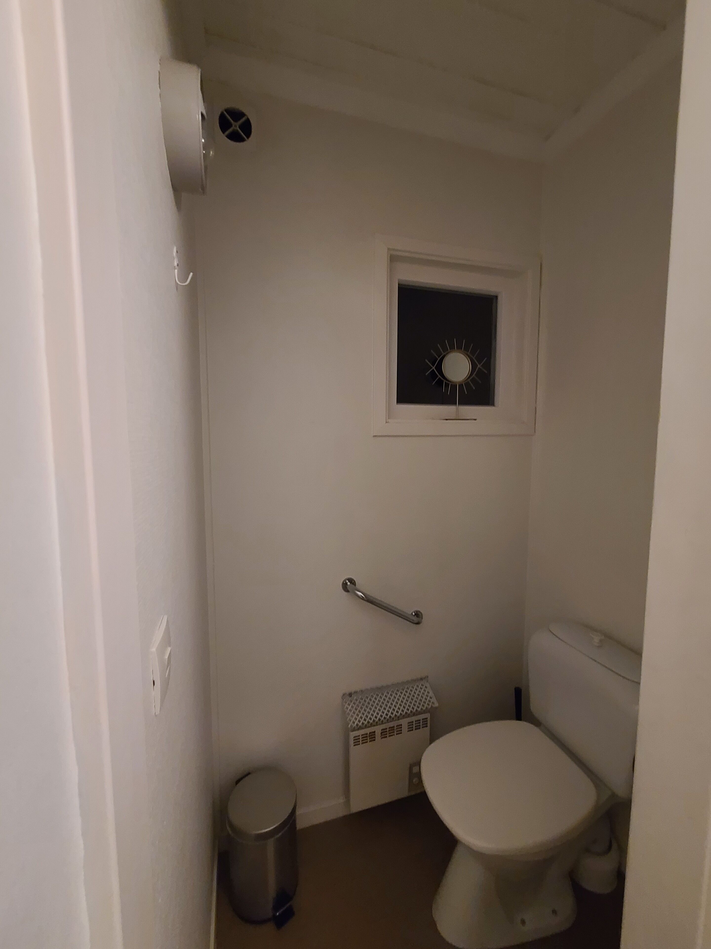 Ett litet, vitt badrum med en toalett, ett stödhandtag, ett fönster och en papperskorg.