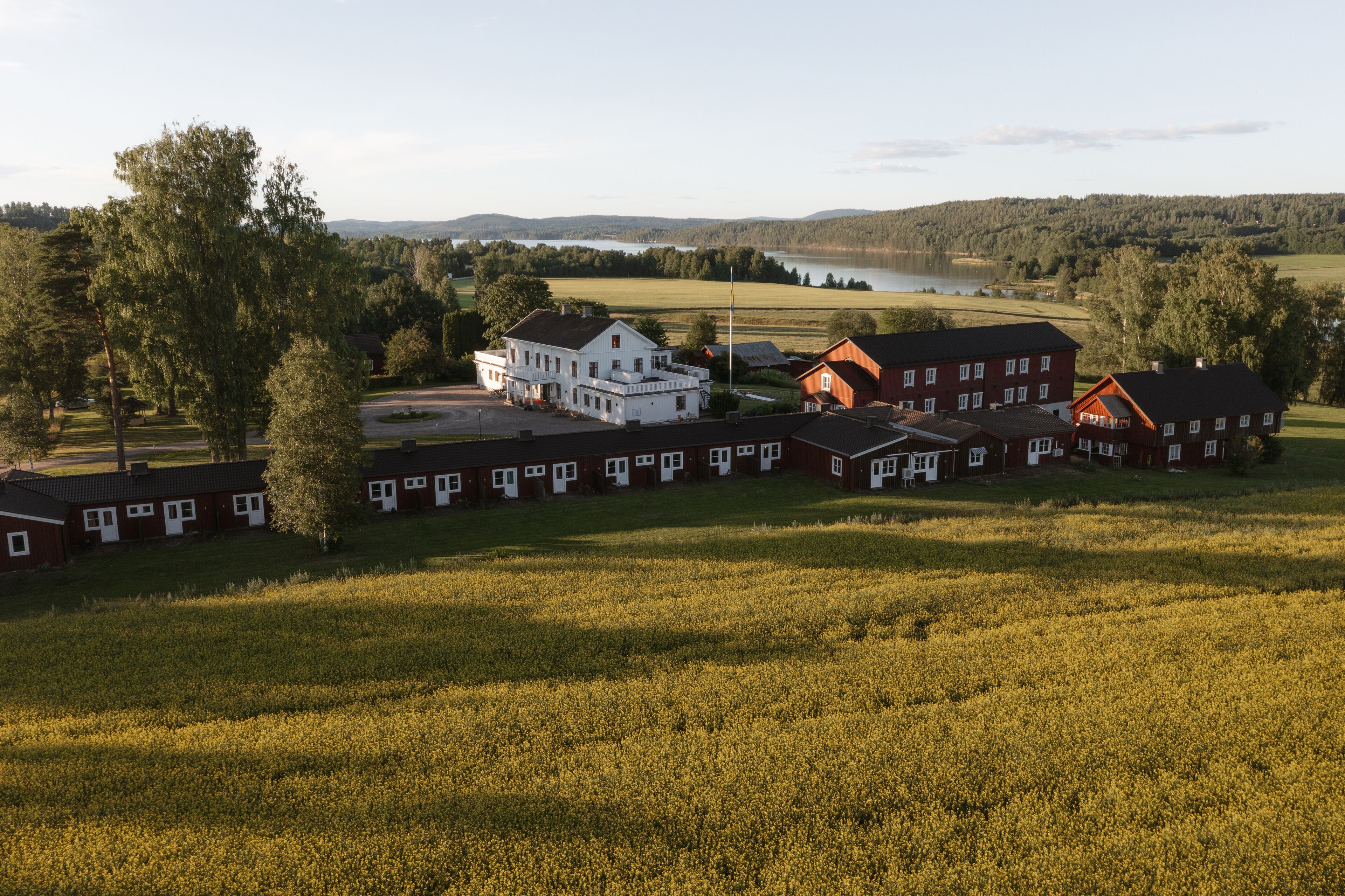 Entorno rural con un edificio blanco y varias casas rojas, rodeado de campos y un lago.