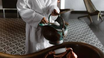 A person in a white robe pouring a liquid from a copper pot into a wooden basin.