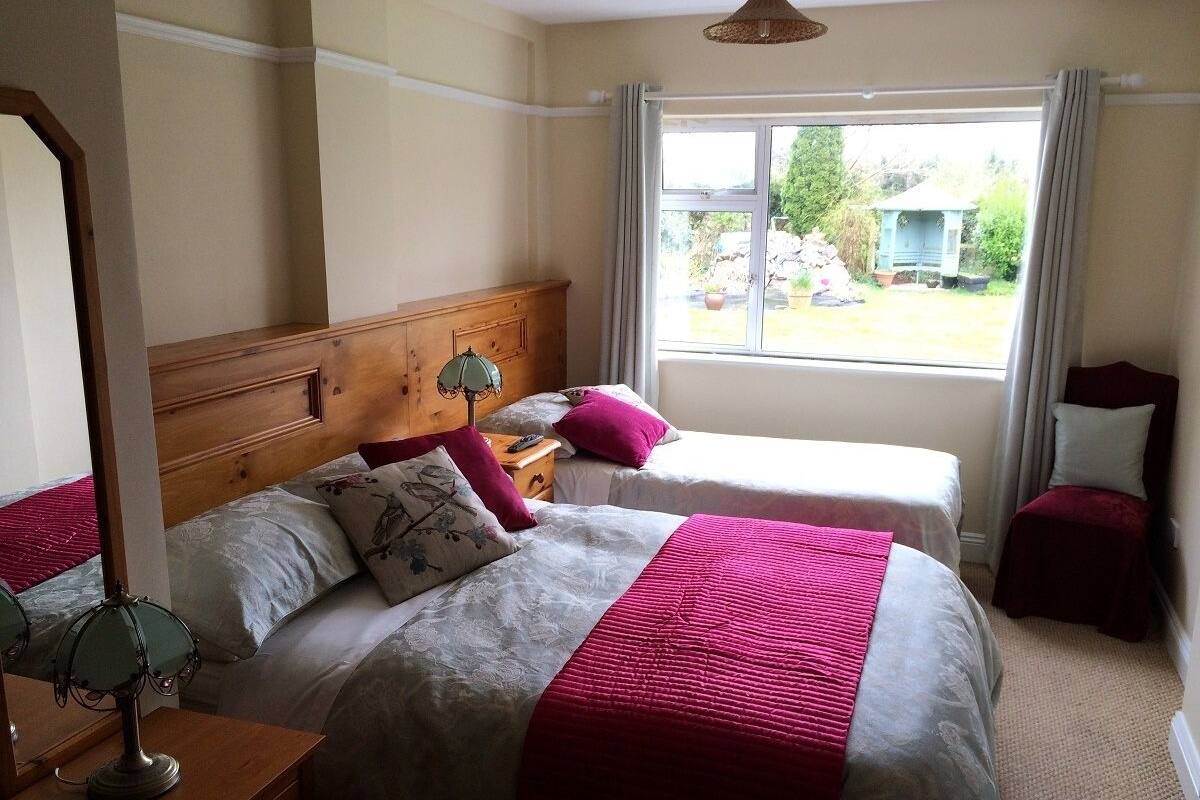 A bedroom with two beds, a wooden headboard, a bedside table with a lamp, and a window with curtains.