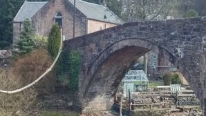 Een stenen brug met een gebogen ontwerp, een bakstenen gebouw met een schuin dak op de achtergrond, en een broeibouw rechts.