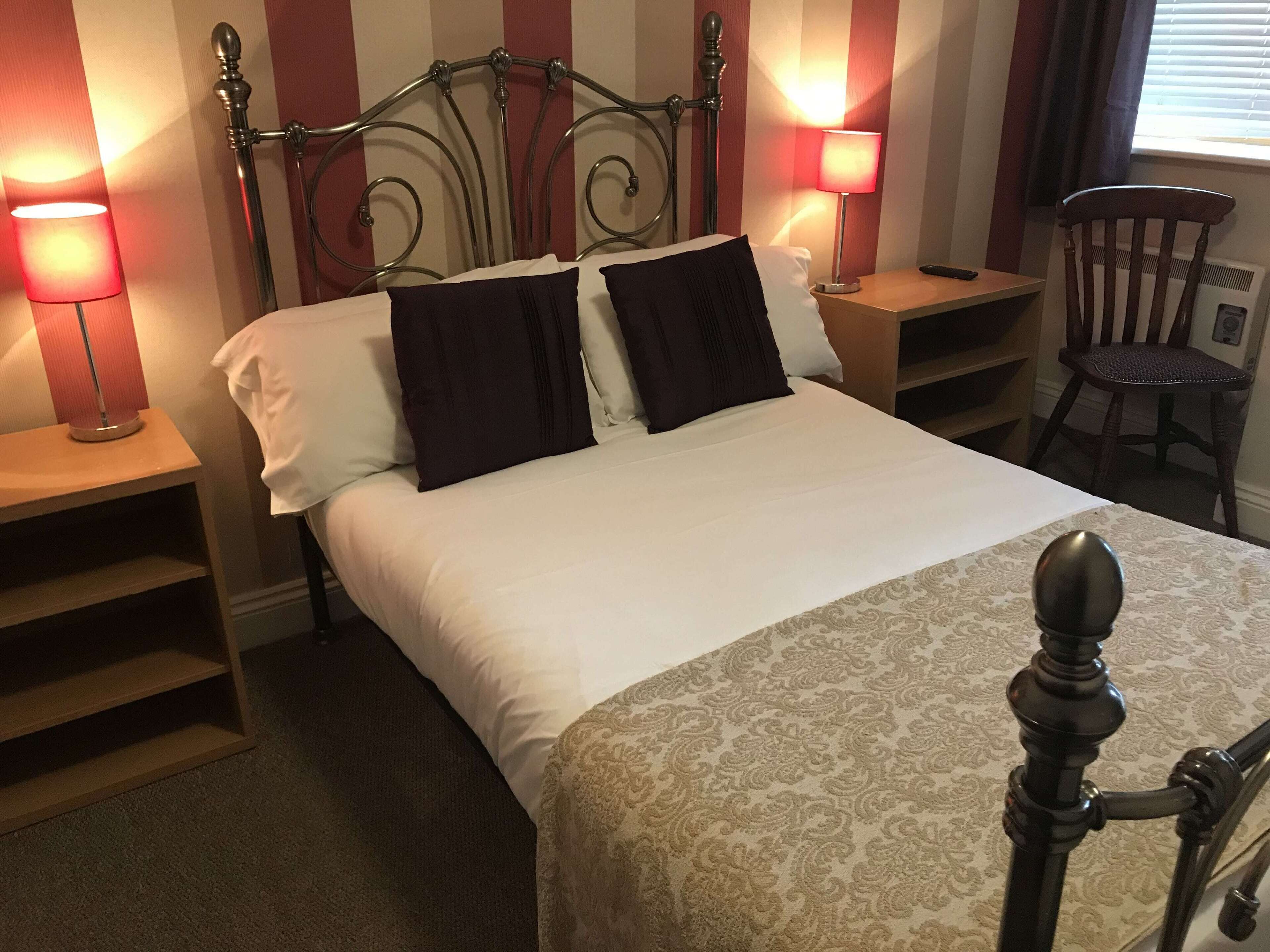 A neatly made bed with a patterned bedspread, two bedside lamps, a wooden chair, and a radiator.