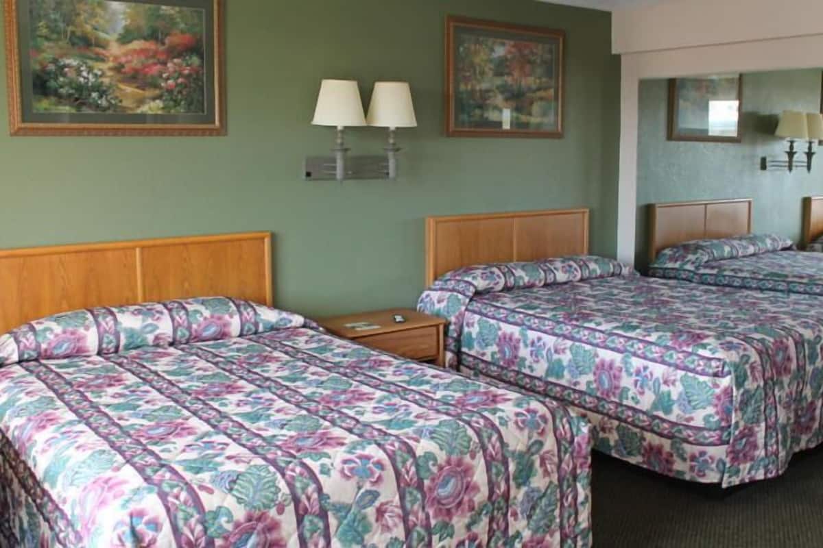 Une chambre d’hôtel avec deux lits, chacun agrémenté de draps à motifs floraux, de têtes de lit en bois et de tables de chevet. Des lampes murales et des œuvres d’art encadrées sont disposées au-dessus des lits.