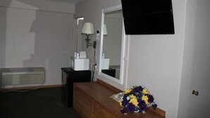 A hotel room with a wooden dresser, a mirror, a flat-screen TV, a lamp, and a heater.
