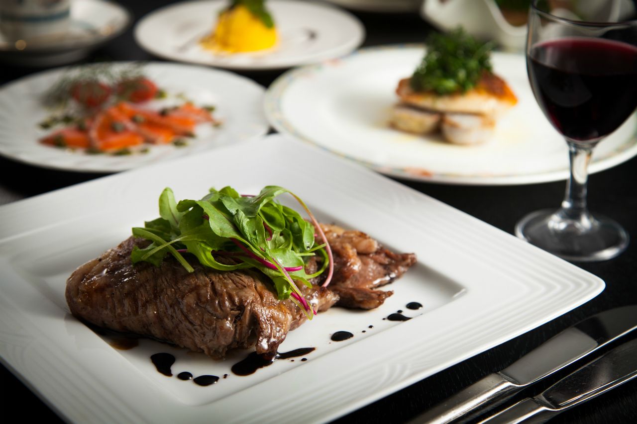 A plate of steak with greens and sauce, a glass of red wine, and other dishes in the background.