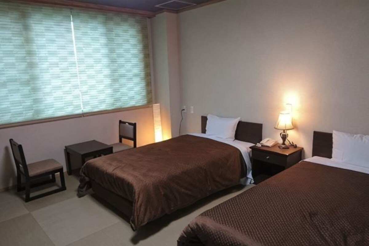 A hotel room with two beds, a desk, a chair, and a window with blinds.