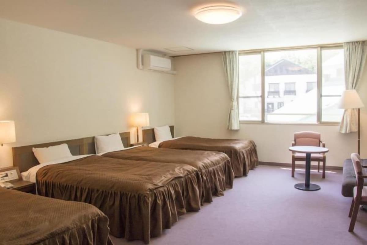 A hotel room with three beds, a round table, a chair, and a window with curtains.