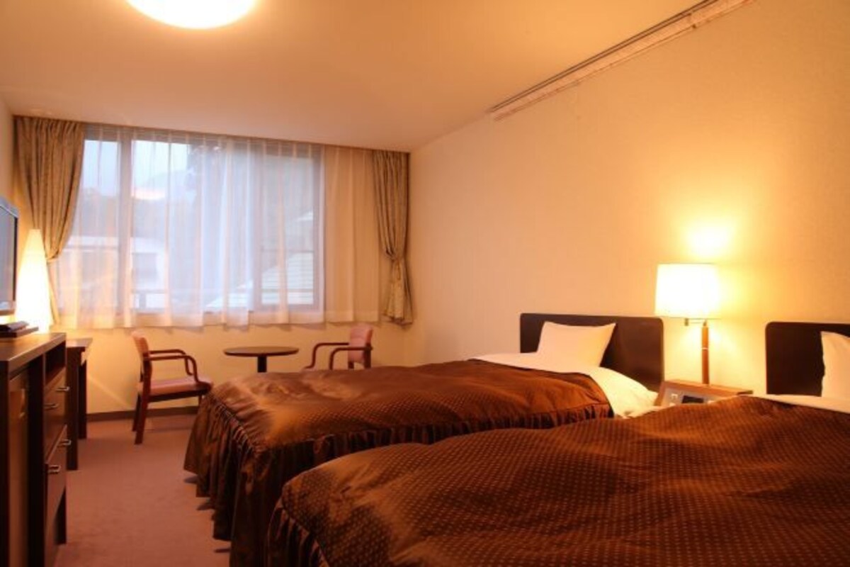 A hotel room with two beds, a desk, a chair, a small table, and a window with curtains.