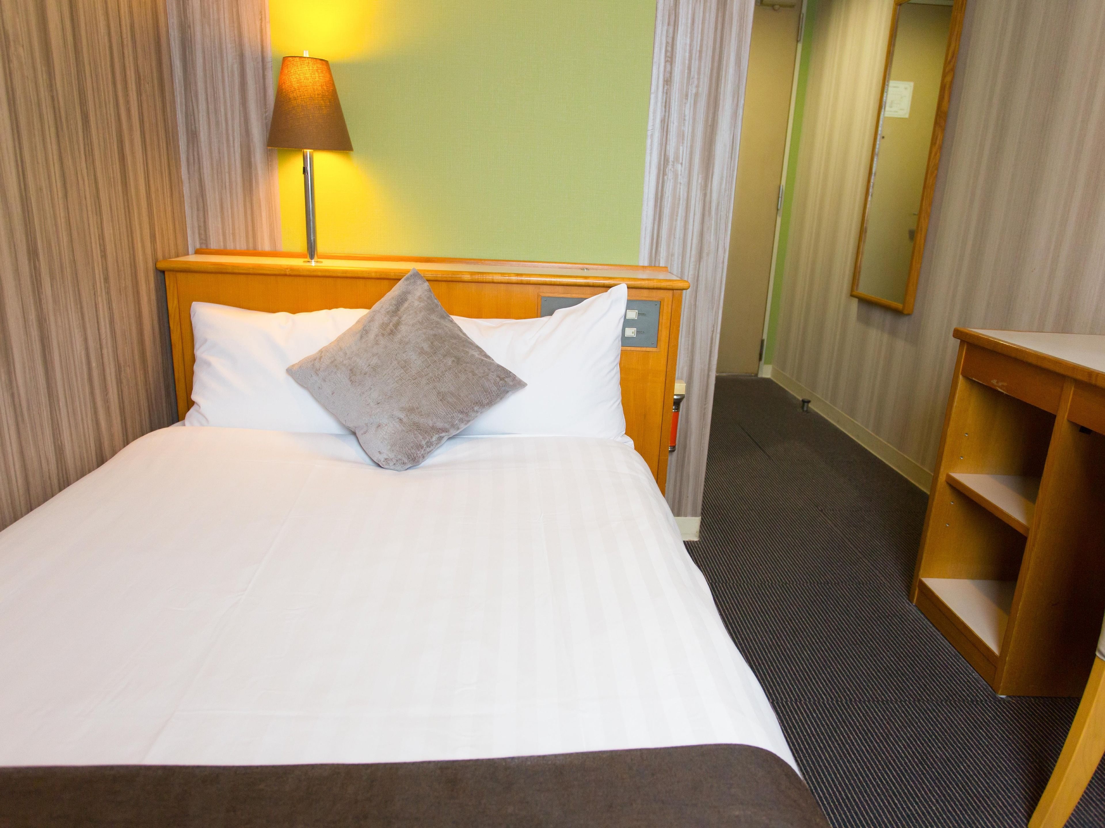 A hotel room with a bed, a nightstand, a lamp, a mirror, and a wooden desk.