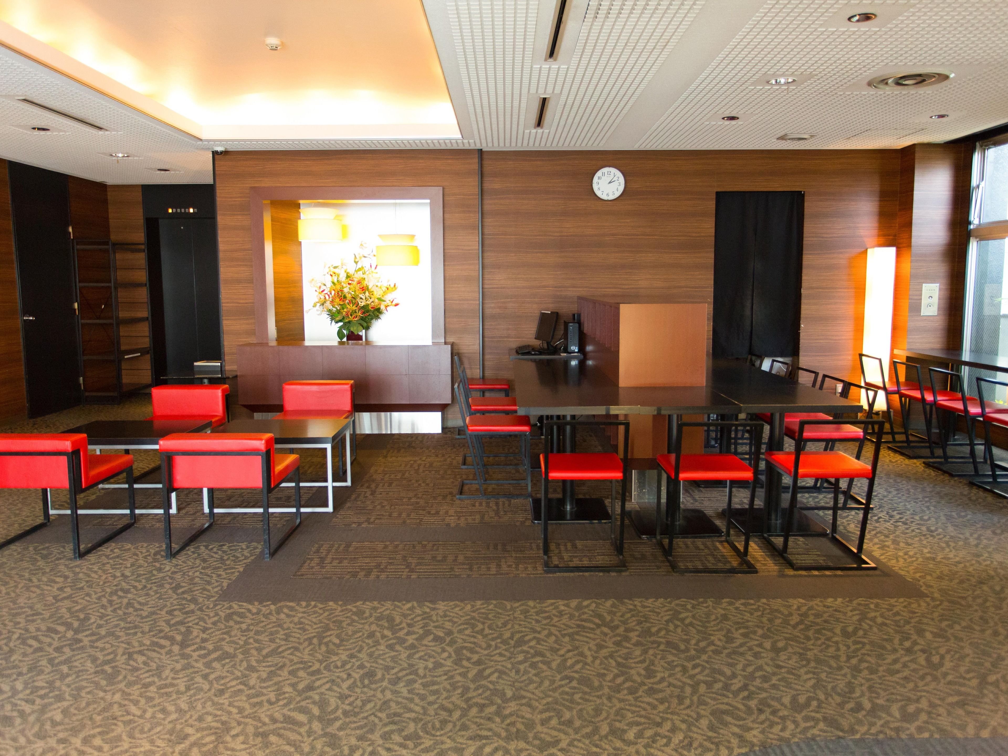 A conference room with a long table, red chairs, a clock on the wall, and a reception desk with a vase of flowers.