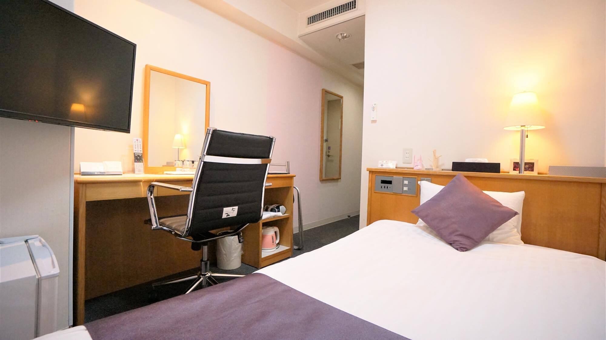 A hotel room with a bed, a desk with a chair, a television, a bedside table with a lamp, and a mirror.
