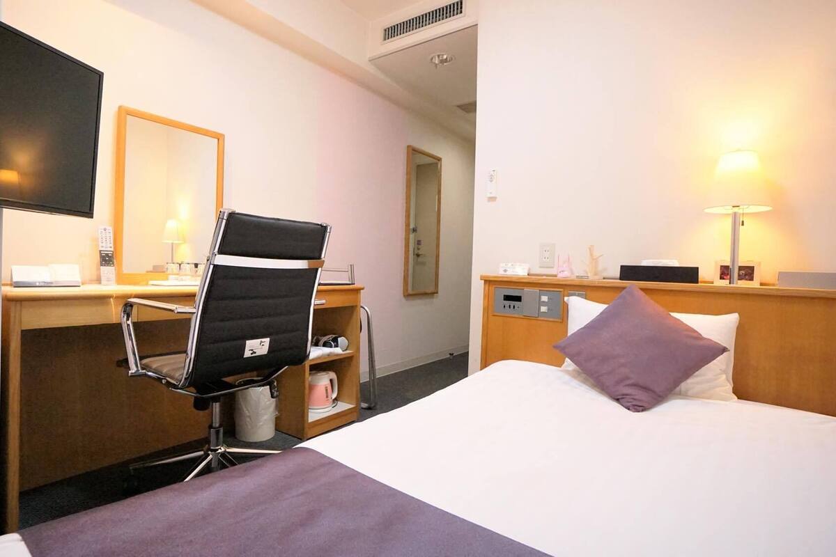 Une chambre d’hôtel comprenant un lit, un bureau avec une chaise, une télévision, une table de chevet avec une lampe et un miroir.