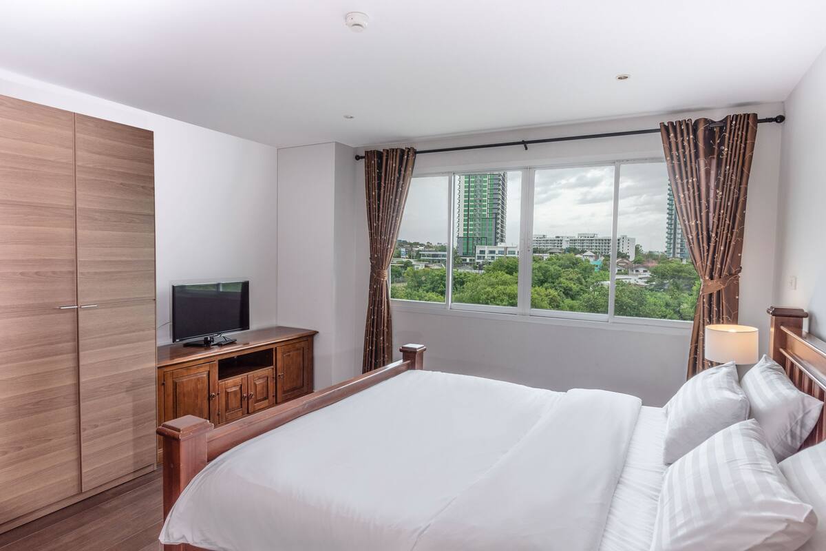 A hotel room with a large bed, a TV on a wooden cabinet, a window with curtains, and a view of the city.