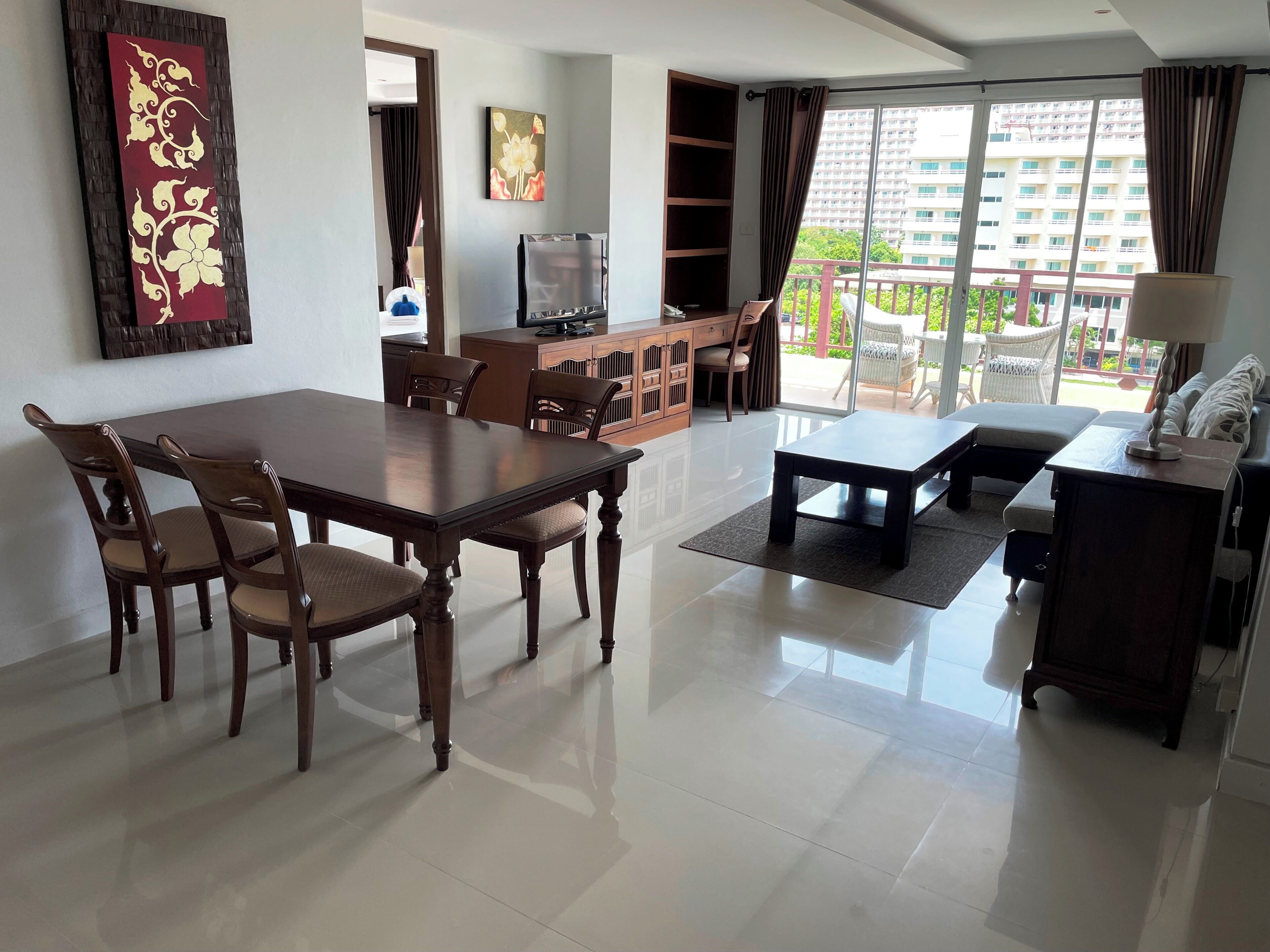 A spacious living room with a dining table, chairs, a sofa, a TV, and a balcony with a view of buildings.
