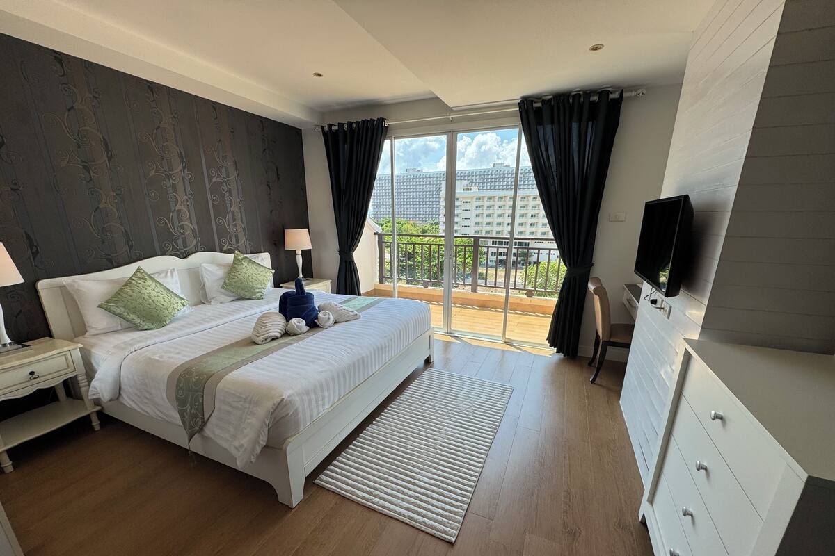 A modern hotel room with a large bed, a TV, a balcony with a view of buildings, and a desk with a lamp.