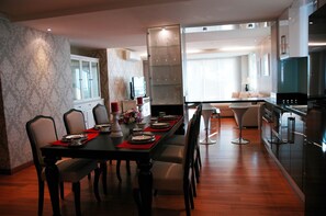 A modern dining area with a long table set for six, a kitchen with stainless steel appliances, and a living area with a sofa and a lamp. - Jomtien Beach Penthouses (Pattaya)