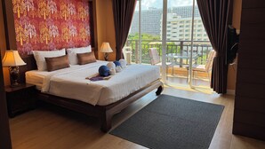 A hotel room with a large bed, a nightstand with a lamp, a view of the city through a balcony with curtains. - Jomtien Beach Penthouses (Pattaya)