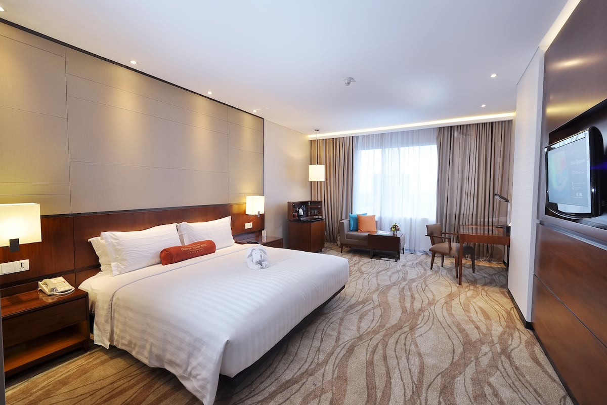 A hotel room with a large bed, bedside tables, a desk, a chair, a TV, and a window with curtains.