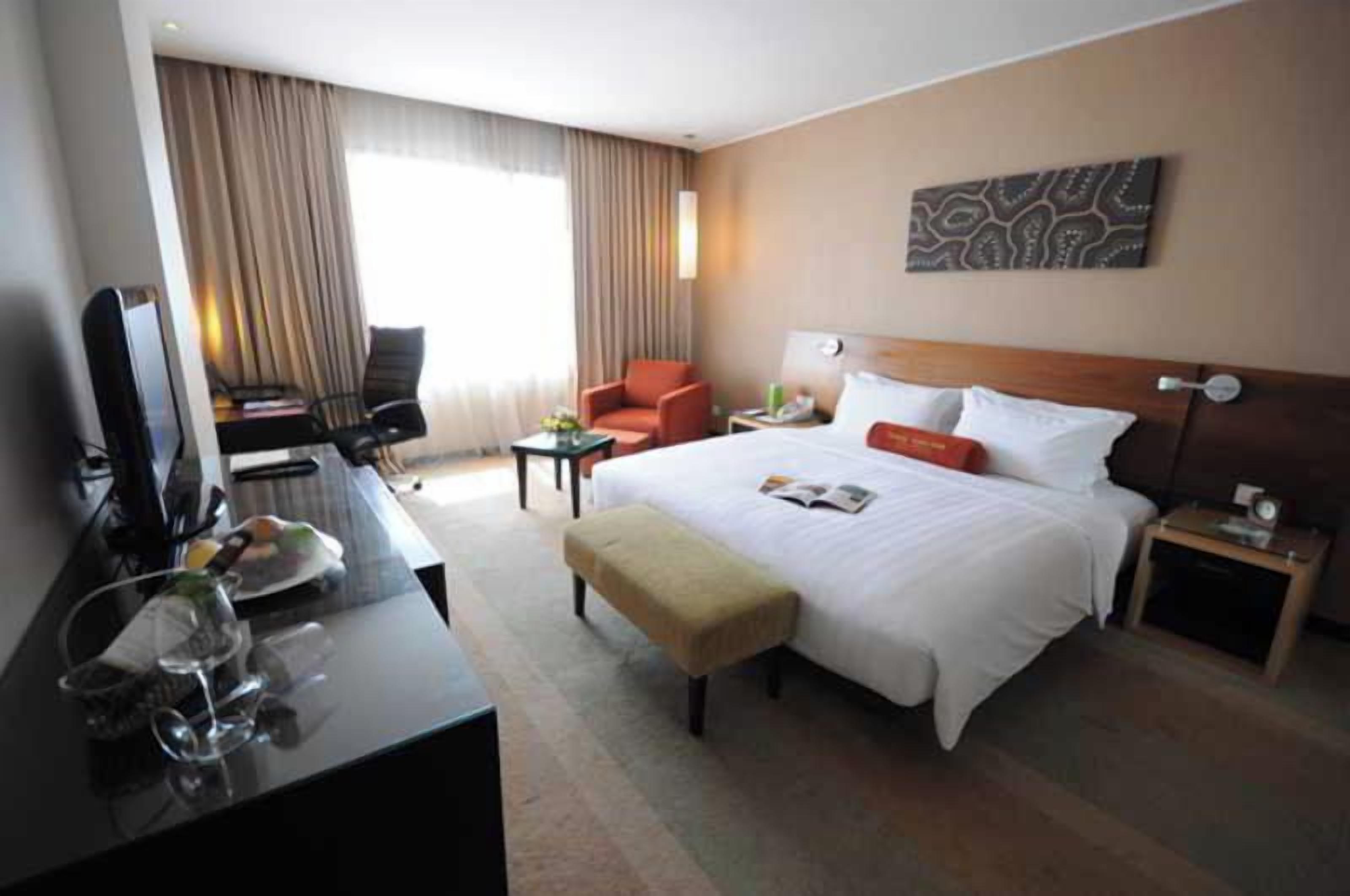 Quarto de hotel com cama, escrivaninha com computador, cadeira, mesinha e mesa de cabeceira.