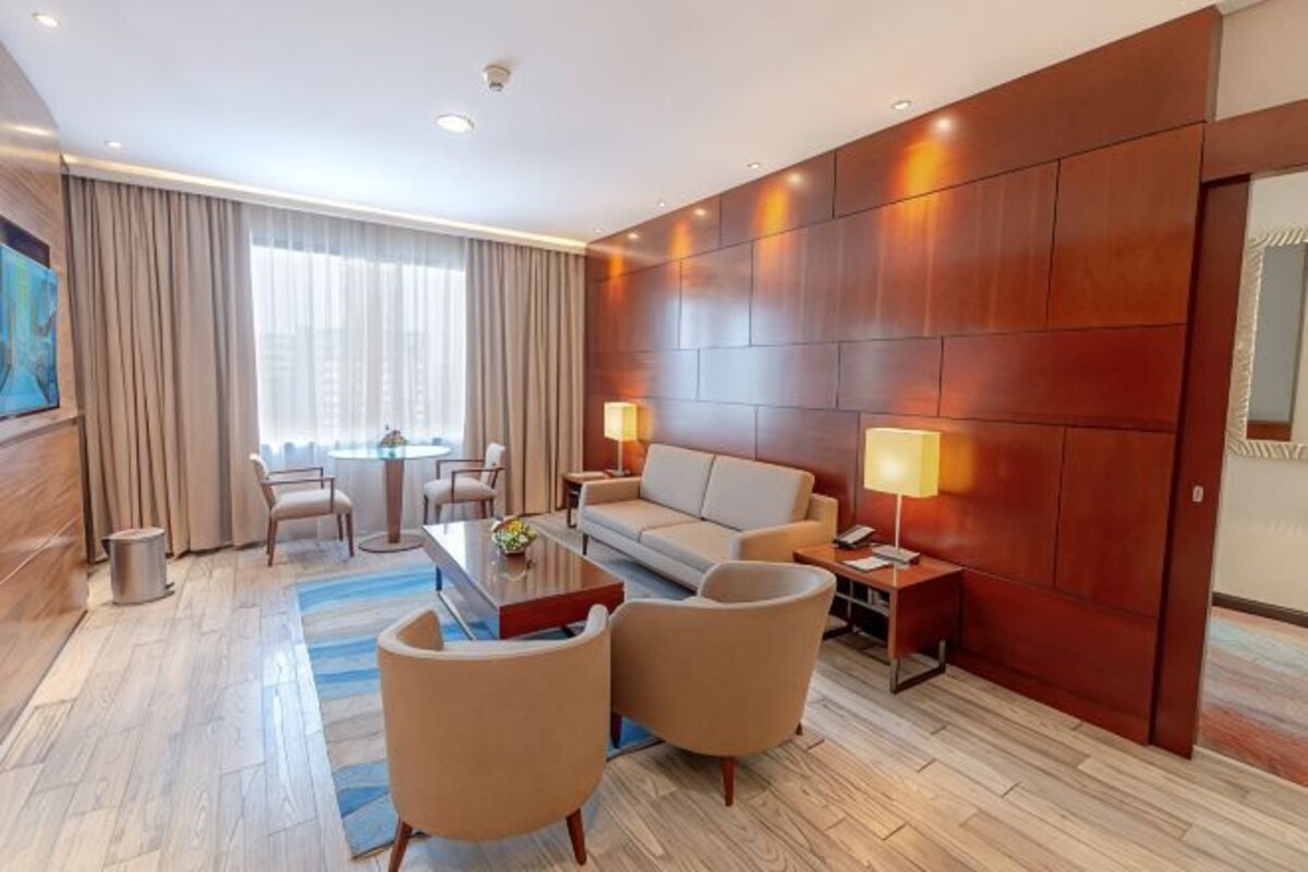 A hotel room with a sofa, two armchairs, a coffee table, a side table with a lamp, a small table with a vase, and a large window with curtains.