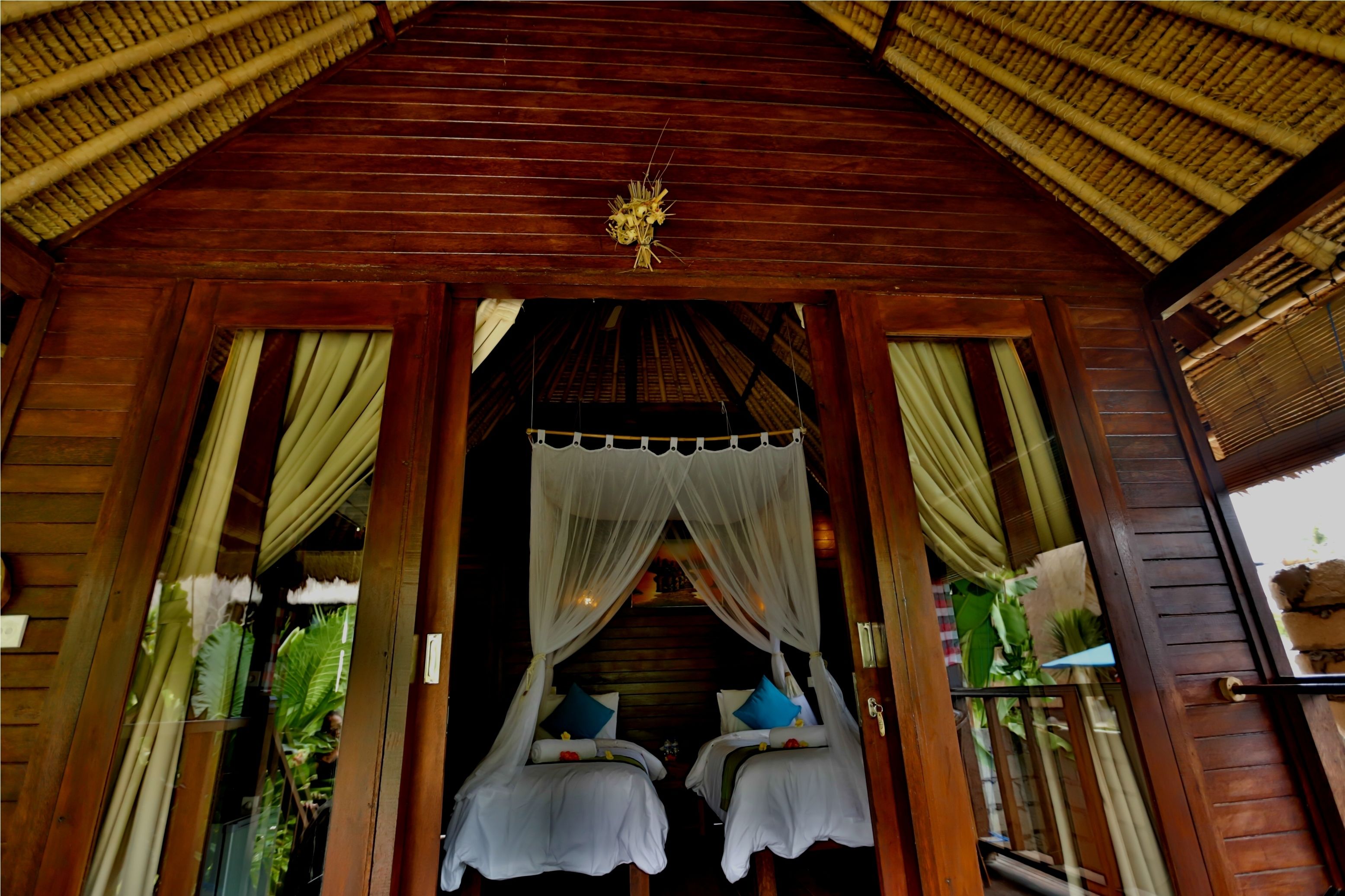 A wooden structure with a thatched roof, two beds with white bedding and blue pillows, and a hanging decorative element above.