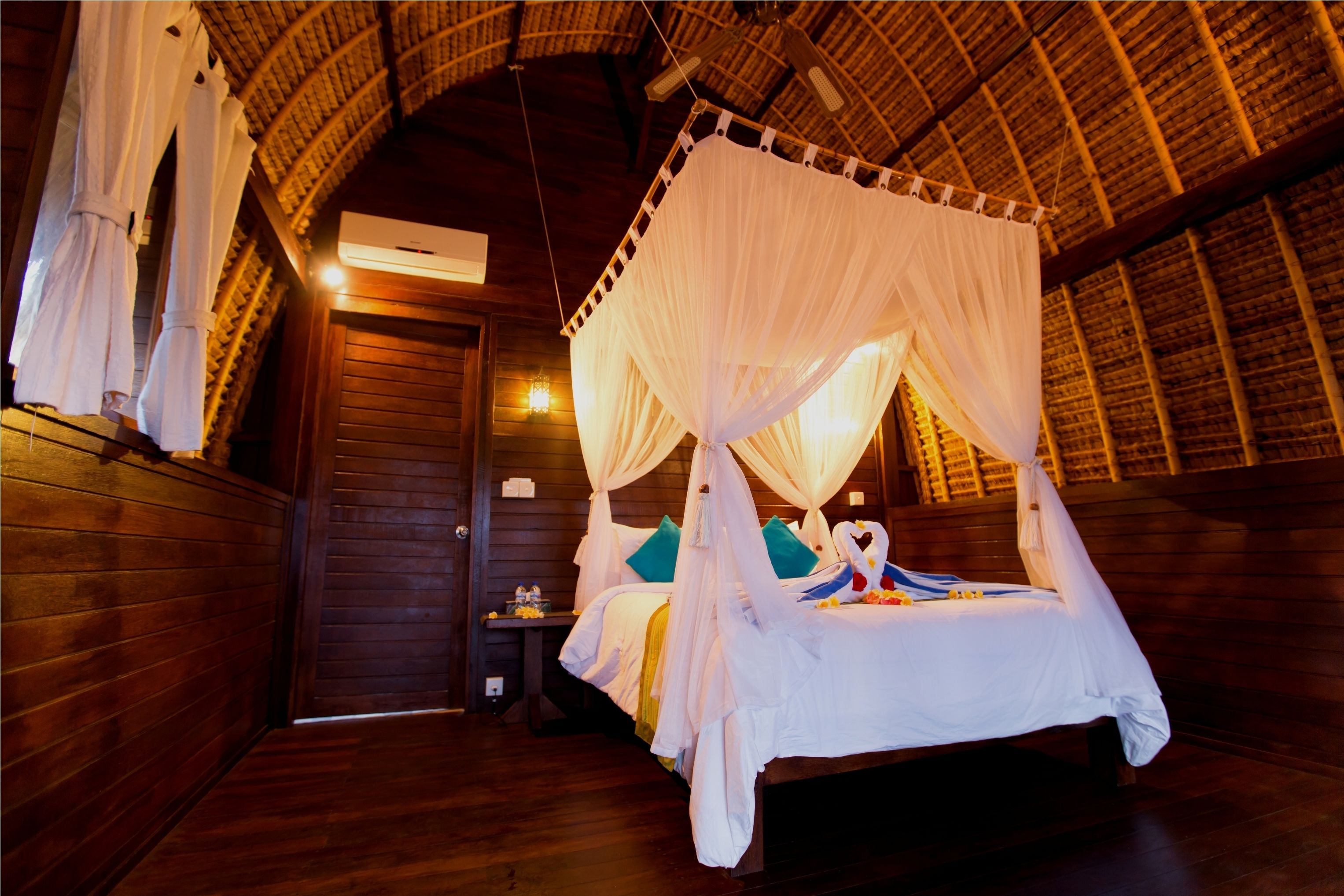 A room with a wooden floor, a bed with white bedding, a canopy bed, and a wooden door.