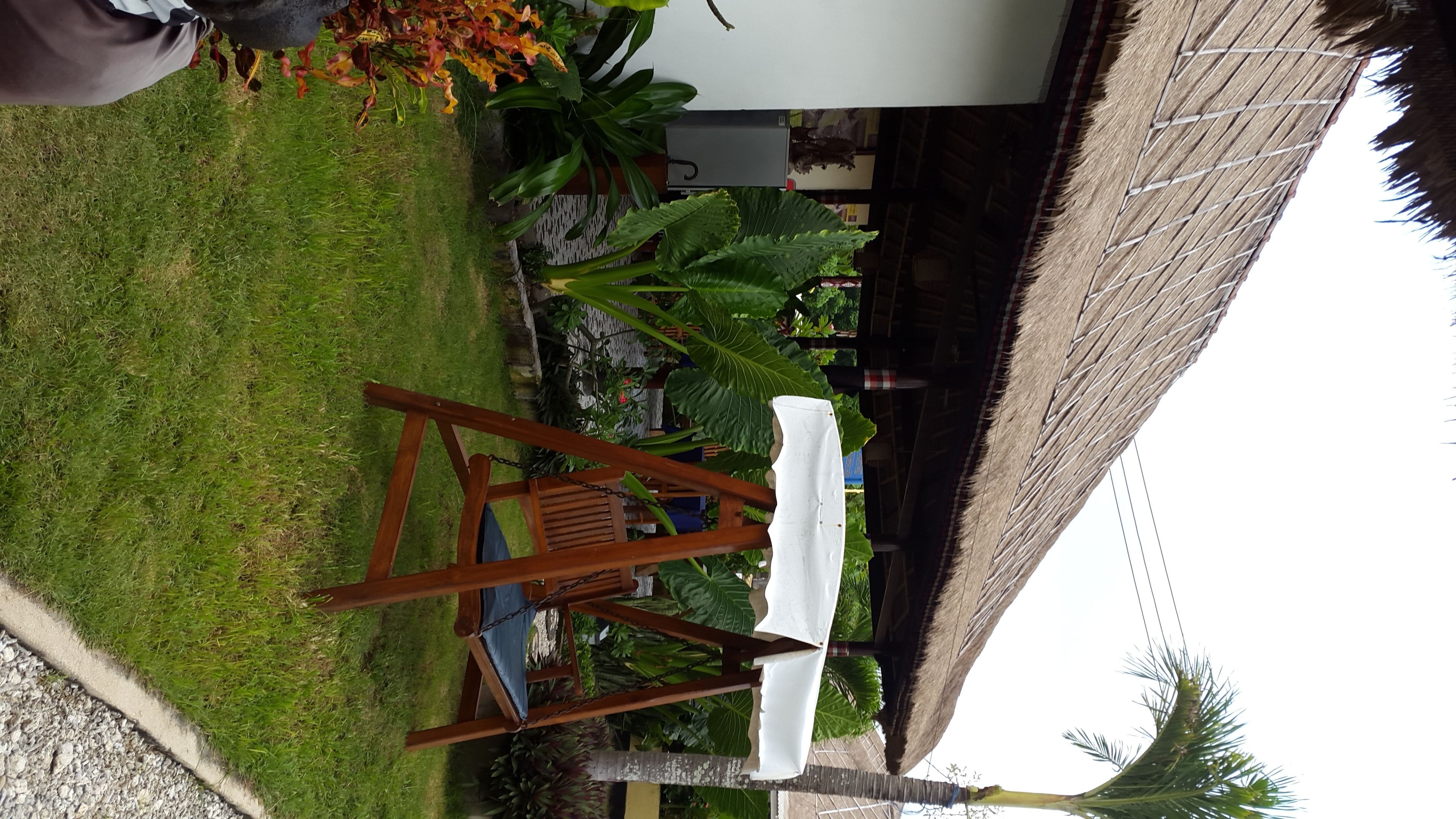 A wooden swing chair with a white cushion in a grassy outdoor area with a thatched roof structure and tropical plants.