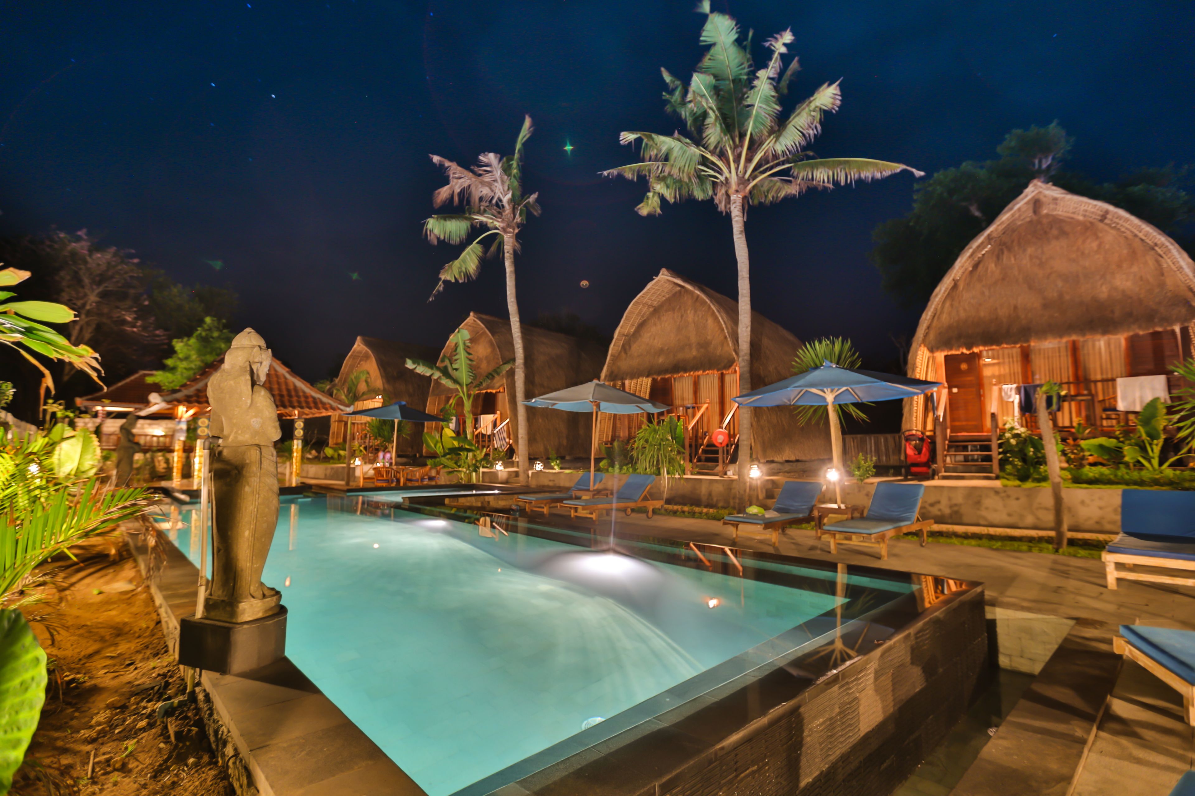 A swimming pool with lounge chairs and umbrellas, surrounded by thatched-roof huts and palm trees.