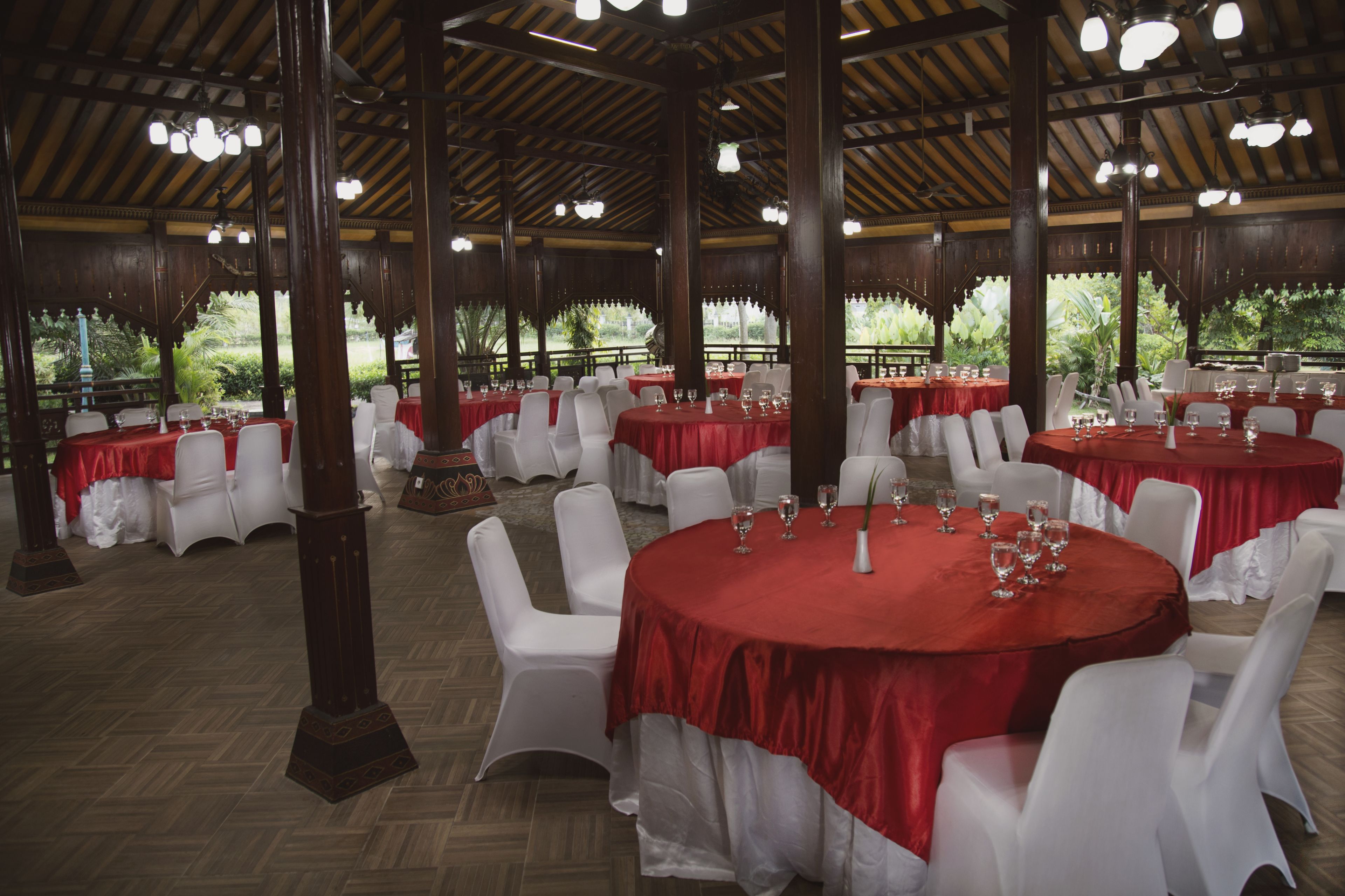 A banquet hall with round tables covered in red tablecloths, white chairs, and wooden pillars.