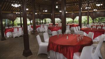 A banquet hall with round tables covered in red tablecloths, white chairs, and wooden pillars.
