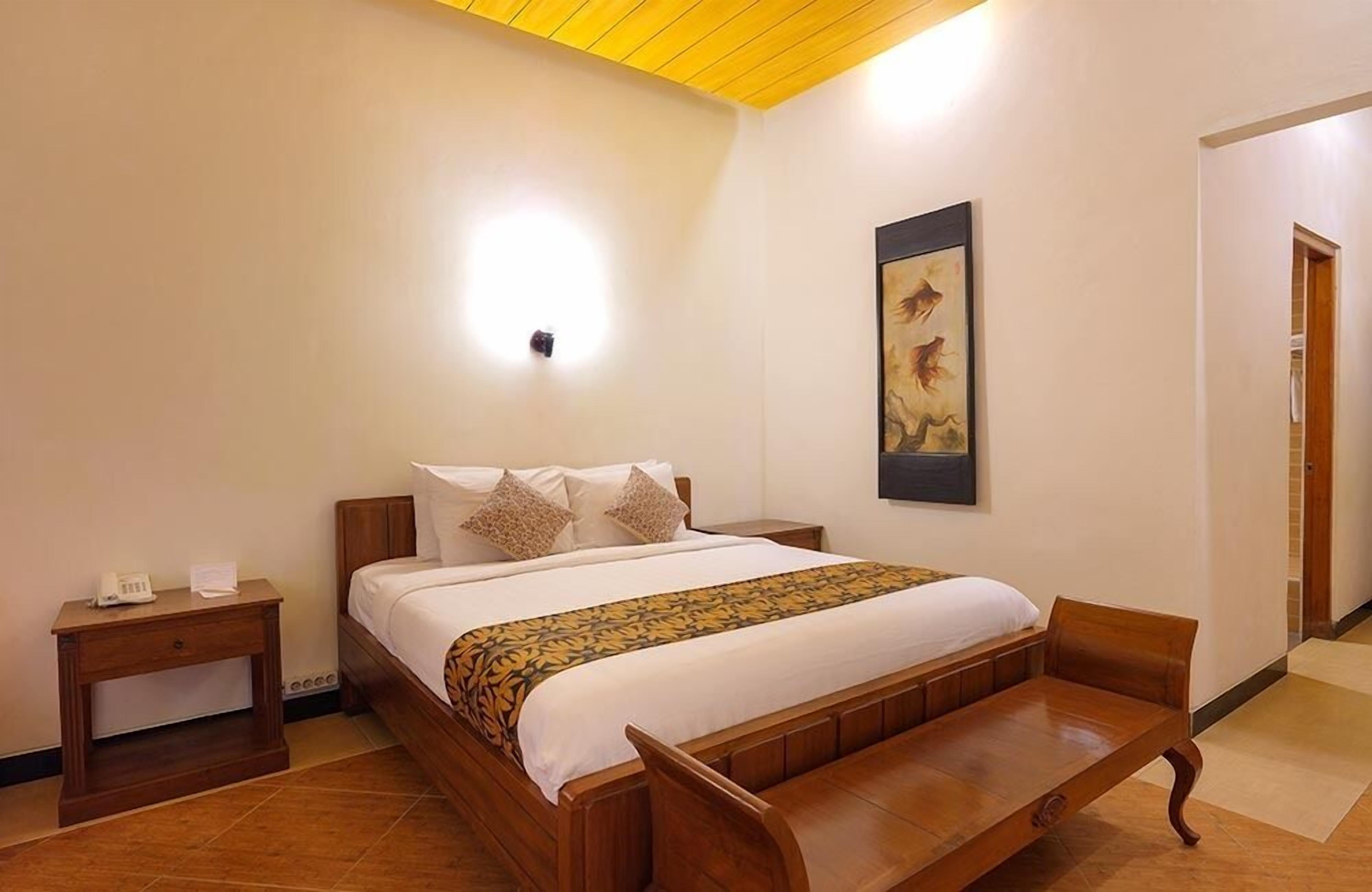 Quarto de hotel com cama de madeira, mesa de cabeceira com telefone, um banco aos pés da cama e um quadro na parede.