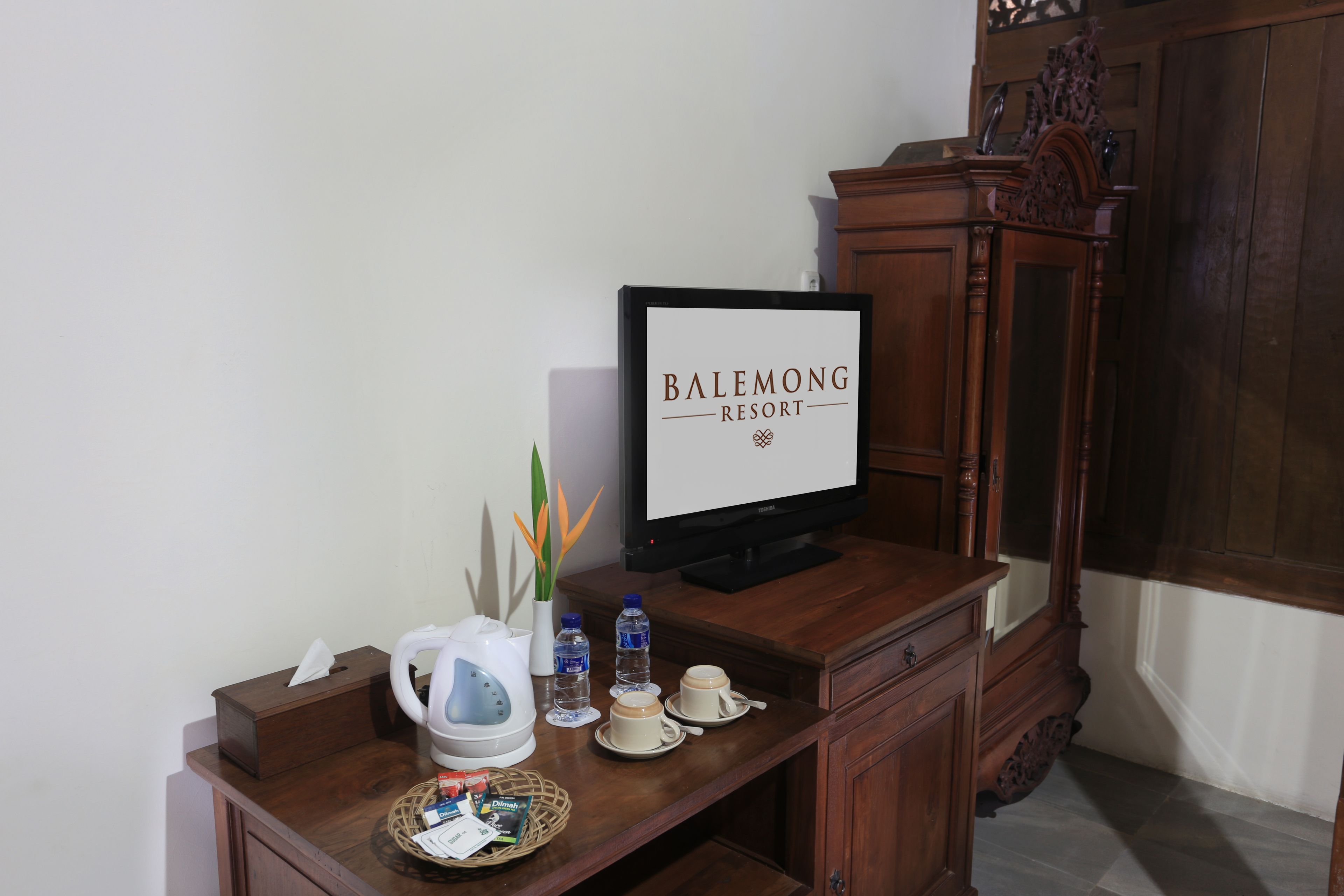 A desk with a television displaying "BALEMONG RESORT", a kettle, water bottles, cups, a tissue box, and a basket with items.