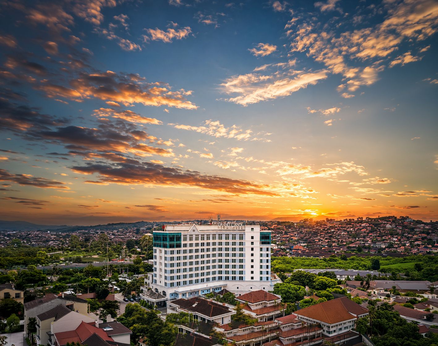 A hotel with a prominent 'GRAND' sign, surrounded by residential buildings and a sunset sky.