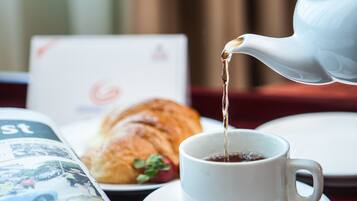A white teapot pouring tea into a white cup on a saucer, a croissant, and a magazine on a table.