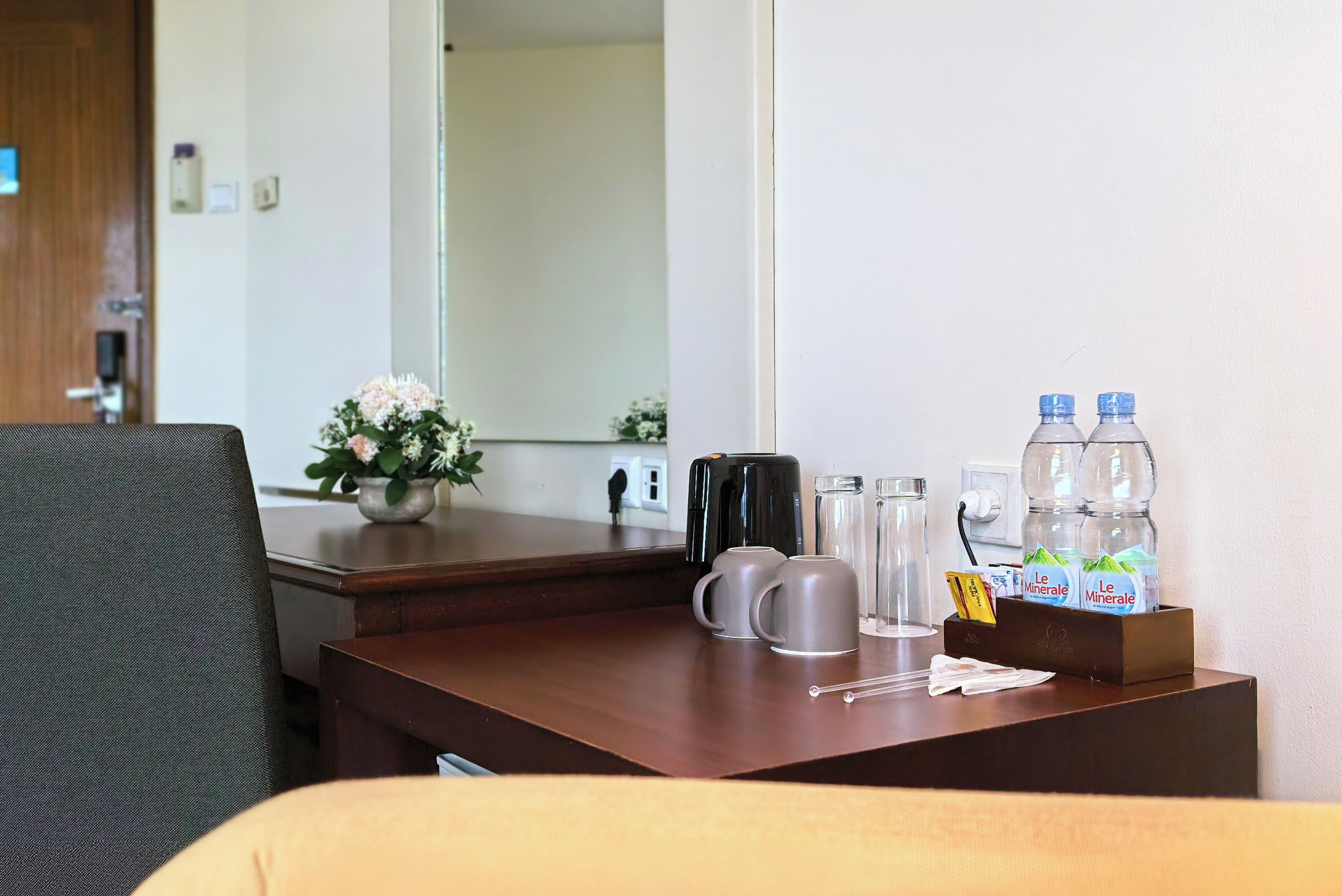 A hotel room with a wooden desk, a coffee maker, two mugs, a glass, a water bottle, and a box of Le Minérale water.