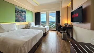 A hotel room with two beds, a desk, a TV, and a large window with a city view.