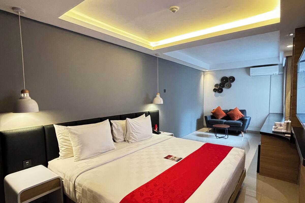 Quarto de hotel moderno com uma cama grande, mesas de cabeceira, escrivaninha e um espaço de estar com sofá e cadeiras.