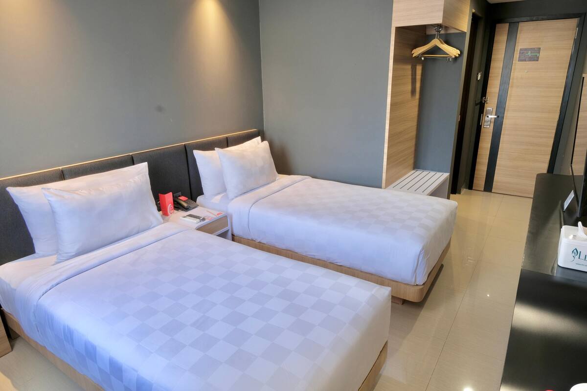 Quarto de hotel com duas camas, uma mesinha de cabeceira com abajur, um cabideiro e uma porta com um aviso.