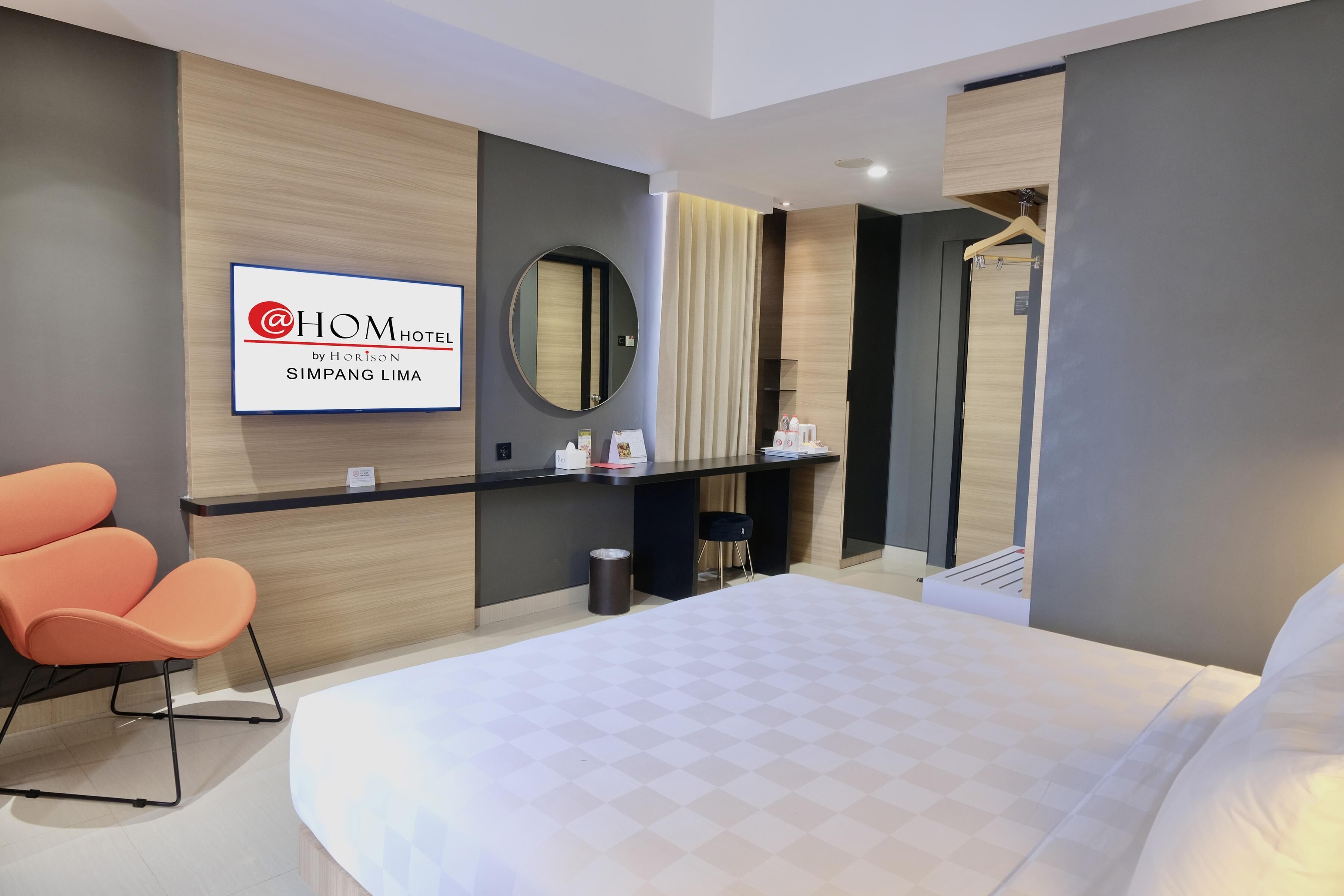 A modern hotel room with a large bed, a desk with a computer, a mirror, and a seating area with orange chairs.