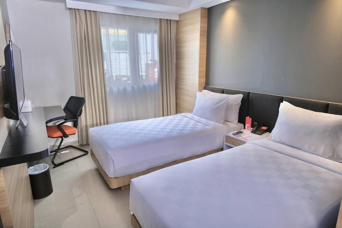 Quarto de hotel com duas camas, uma escrivaninha, uma cadeira, uma televisão e uma janela com cortinas.