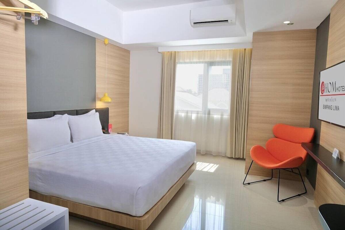 Quarto de hotel com cama, escrivaninha, cadeira, janela com cortinas e um aviso na parede.