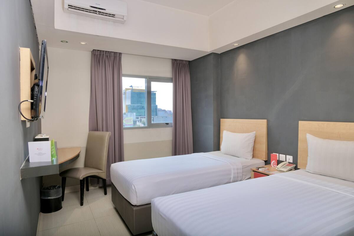 Quarto de hotel com duas camas, uma escrivaninha, uma cadeira, um telefone e uma janela com cortinas.