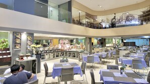 A multi-level restaurant with checkered tablecloths, a central bar area, and a view of a cityscape through large windows. - @HOM Simpang Lima Semarang (Semarang)