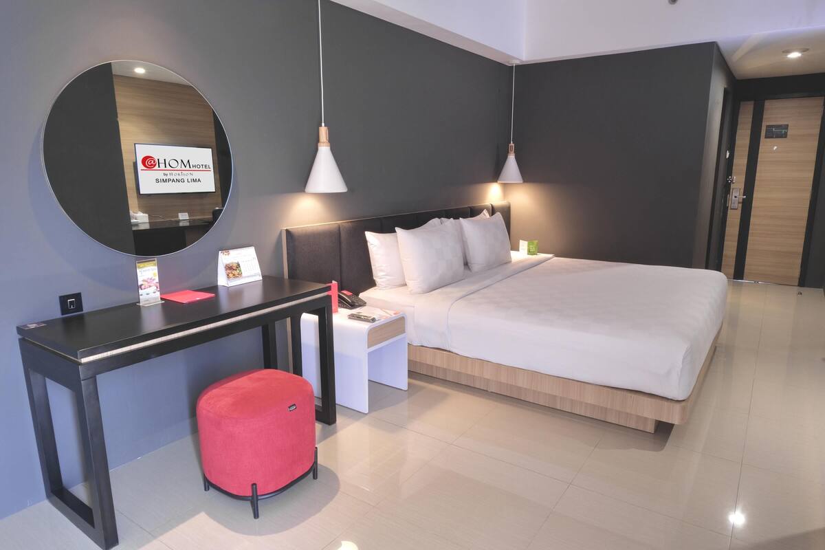 Quarto de hotel moderno com uma cama grande, uma escrivaninha com cadeira, um espelho redondo e duas luminárias pendentes.