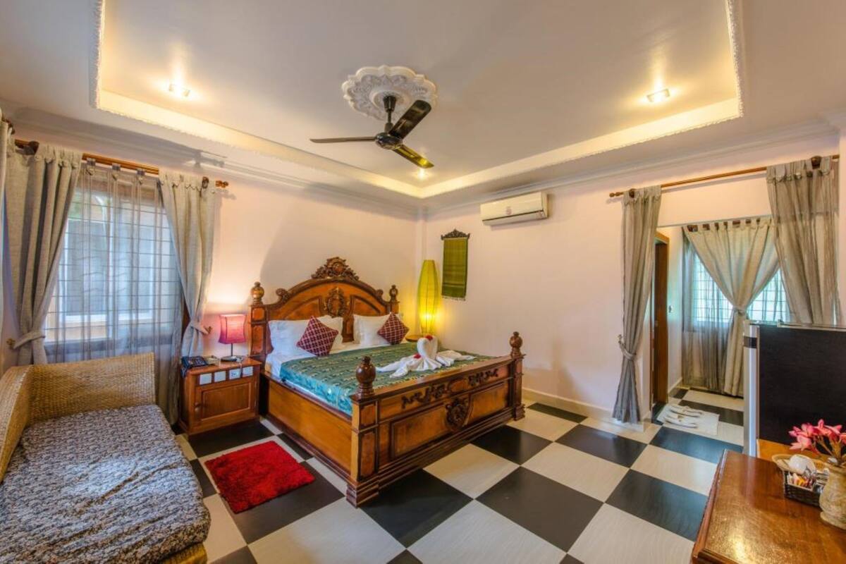 Un dormitorio con suelo a cuadros, una cama de madera con cabecera decorativa, una mesita de noche y una lámpara en la mesilla, además de un ventilador de techo.