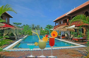 A pool area with lounge chairs and umbrellas, a thatched-roof structure, and a building with a tiled roof. - Delux Villa (Battambang)