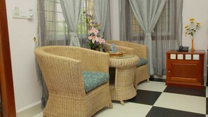 Una habitación con dos sillas de mimbre, una mesita redonda con una bandeja, un florero con flores y un armario de madera que contiene un teléfono y una botella de agua.
