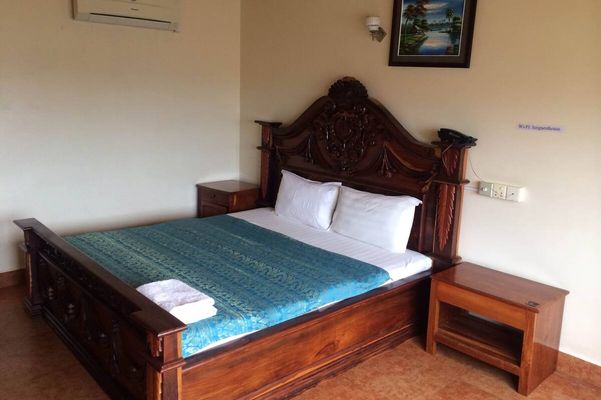 Una cama de madera con almohadas blancas, una colcha azul y una mesita de noche con una lámpara.