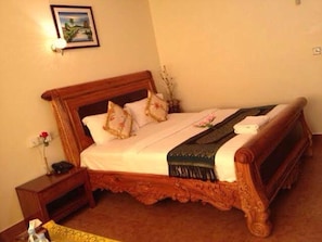 Una cama de madera con colcha blanca y almohadillas decorativas, una mesita de noche con un florero y un cuadro en la pared.